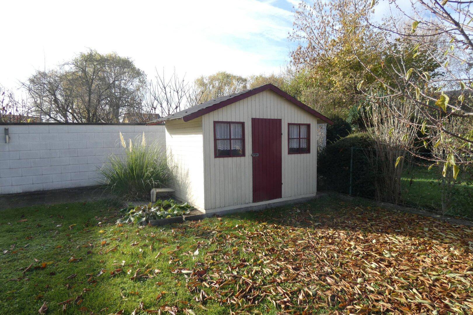 Kleines, charmantes Gartenhaus mit roter Tür und Fenstern, umgeben von Herbstlaub.