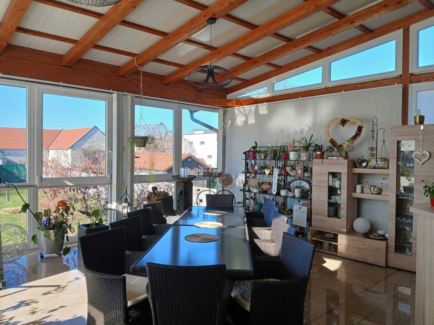 Helles Esszimmer in einem Wintergarten mit großen Fenstern, Holzbalken und Blick ins Grüne.