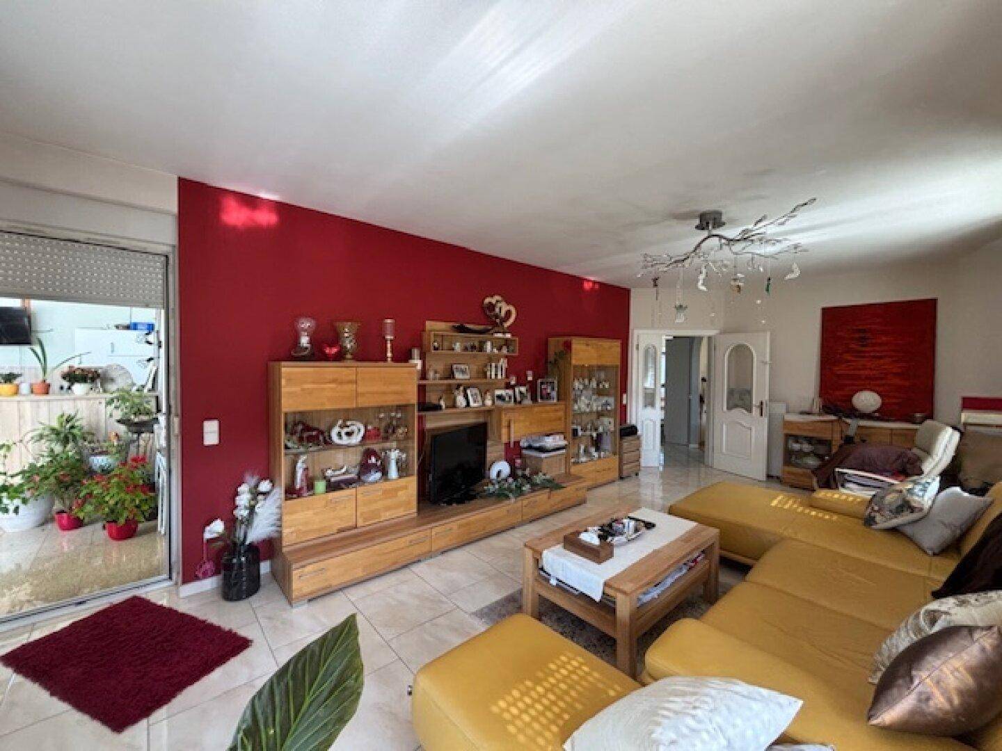 Geräumiges Wohnzimmer mit roter Akzentwand, bequemer Couch und Zugang zu einem verglasten Bereich.