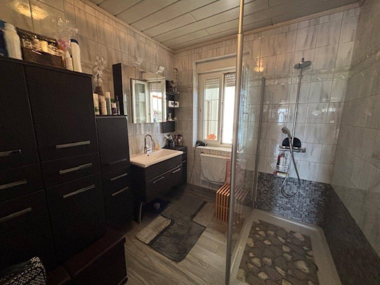 Modernes Badezimmer mit Duschkabine, Waschbecken, dunklen Möbeln und einem Fenster für Tageslicht.