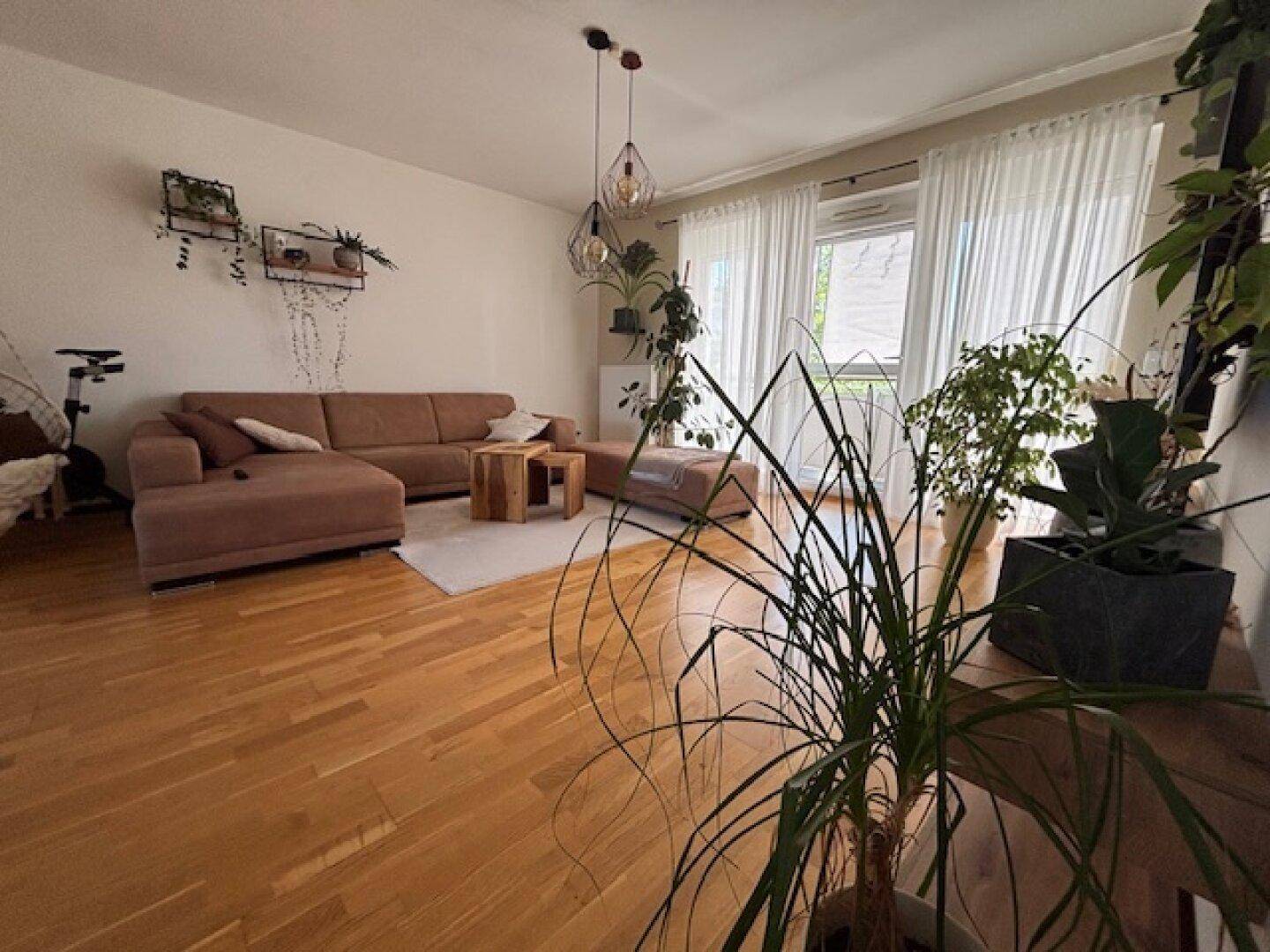 Gemütlicher Wohnbereich mit beiger Couch, Holzboden und großen Fenstern, die viel Tageslicht hereinlassen.
