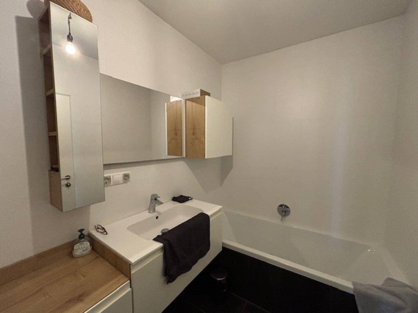 Badezimmer mit Badewanne, Einzelwaschbecken und Spiegelschrank, ausgestattet mit dunklen Bodenfliesen.