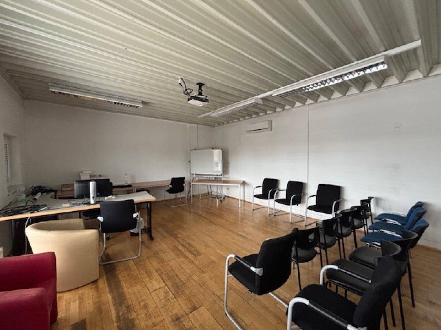 Konferenz- oder Schulungsraum mit Holzfußboden, Beamer, Whiteboard und Reihen von Stühlen, unter einer Wellblechdecke.