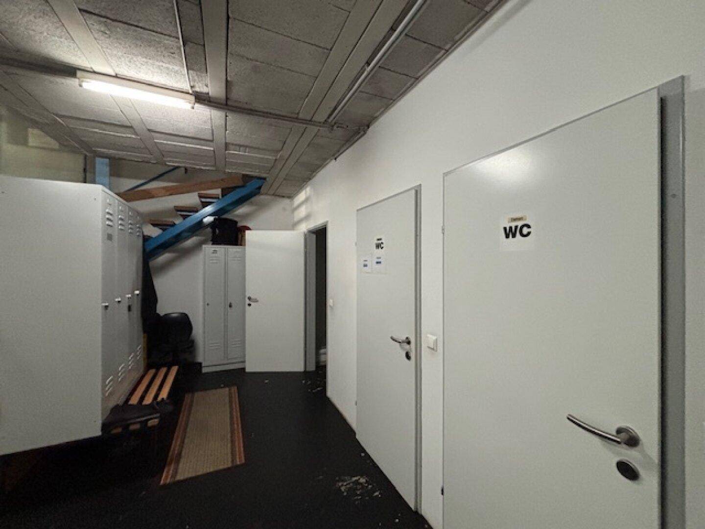 Flur mit mehreren Türen, darunter Toiletten, und Spinden auf der linken Seite, unter einer unverkleideten Decke.