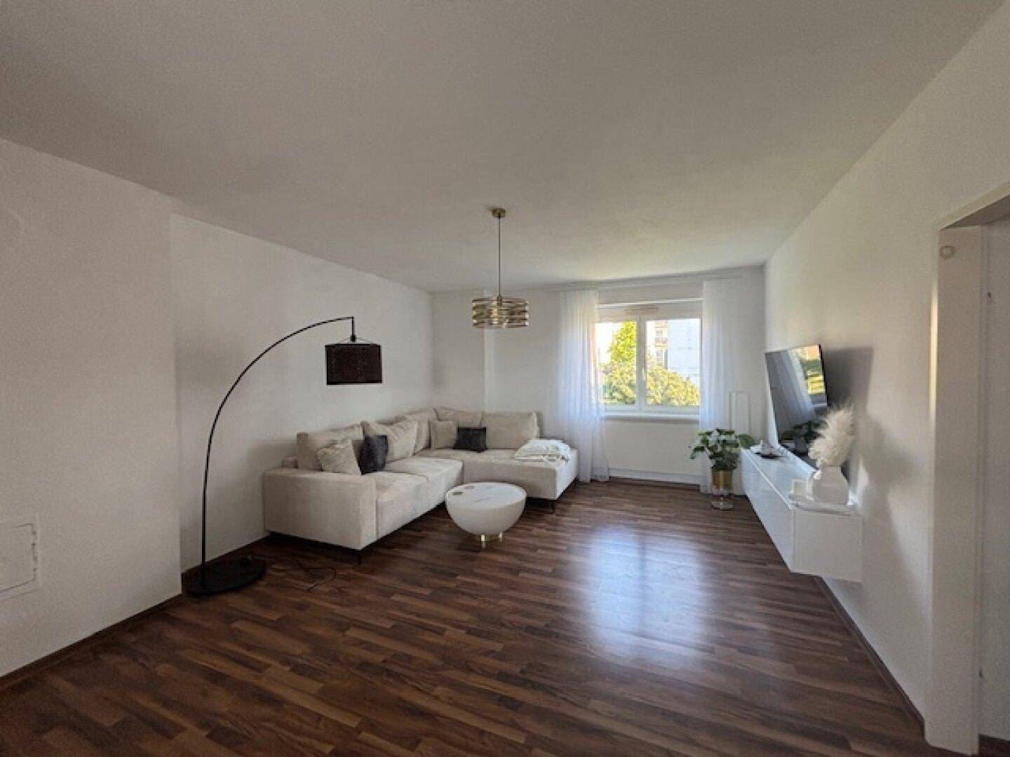 Geräumiges Wohnzimmer mit beigem Ecksofa, Holzboden und Blick ins Grüne.
