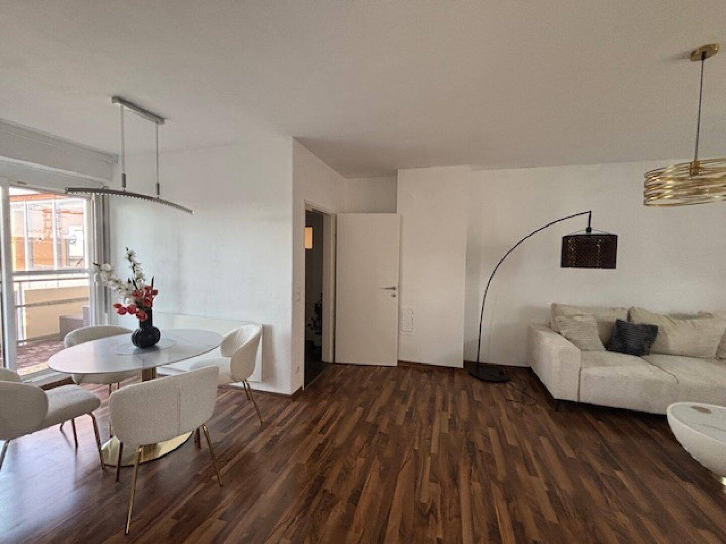 Offener Wohn- und Essbereich mit Holzboden, Sofa und Zugang zum Balkon.