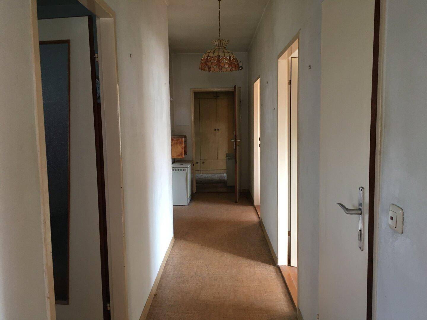 Langer Flur mit Teppichboden und mehreren Türen, beleuchtet von einer Deckenlampe.