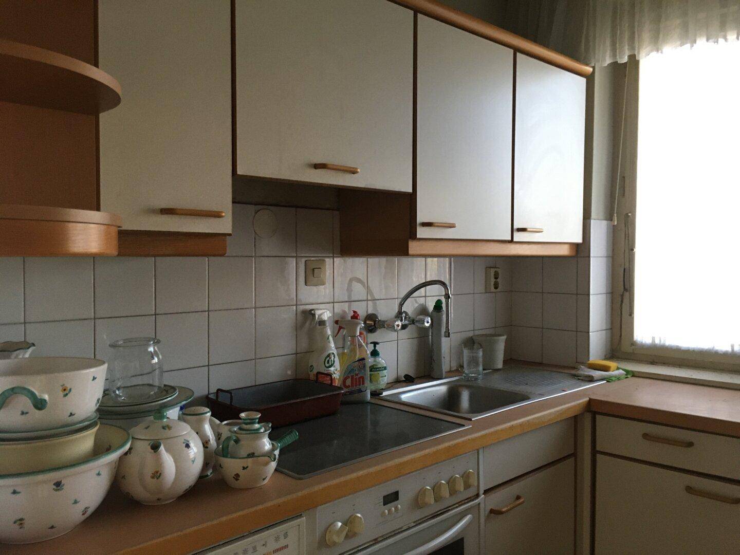 Einfache Küche mit weißen Hängeschränken, Spüle und Herd, in die Jahre gekommen.