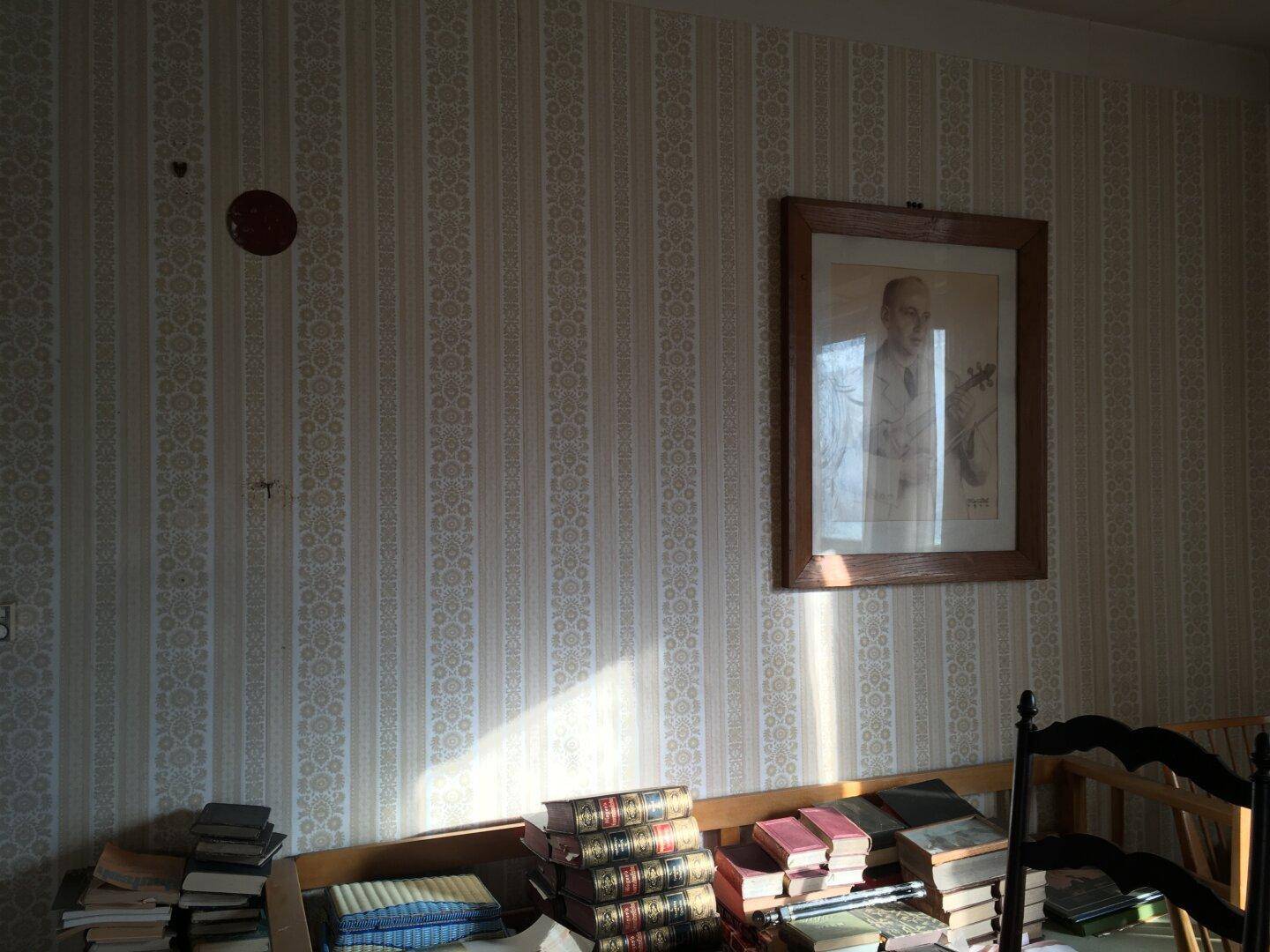 Raum mit gemusterter Tapete und einem Bücherregal, beleuchtet durch Sonnenlicht.