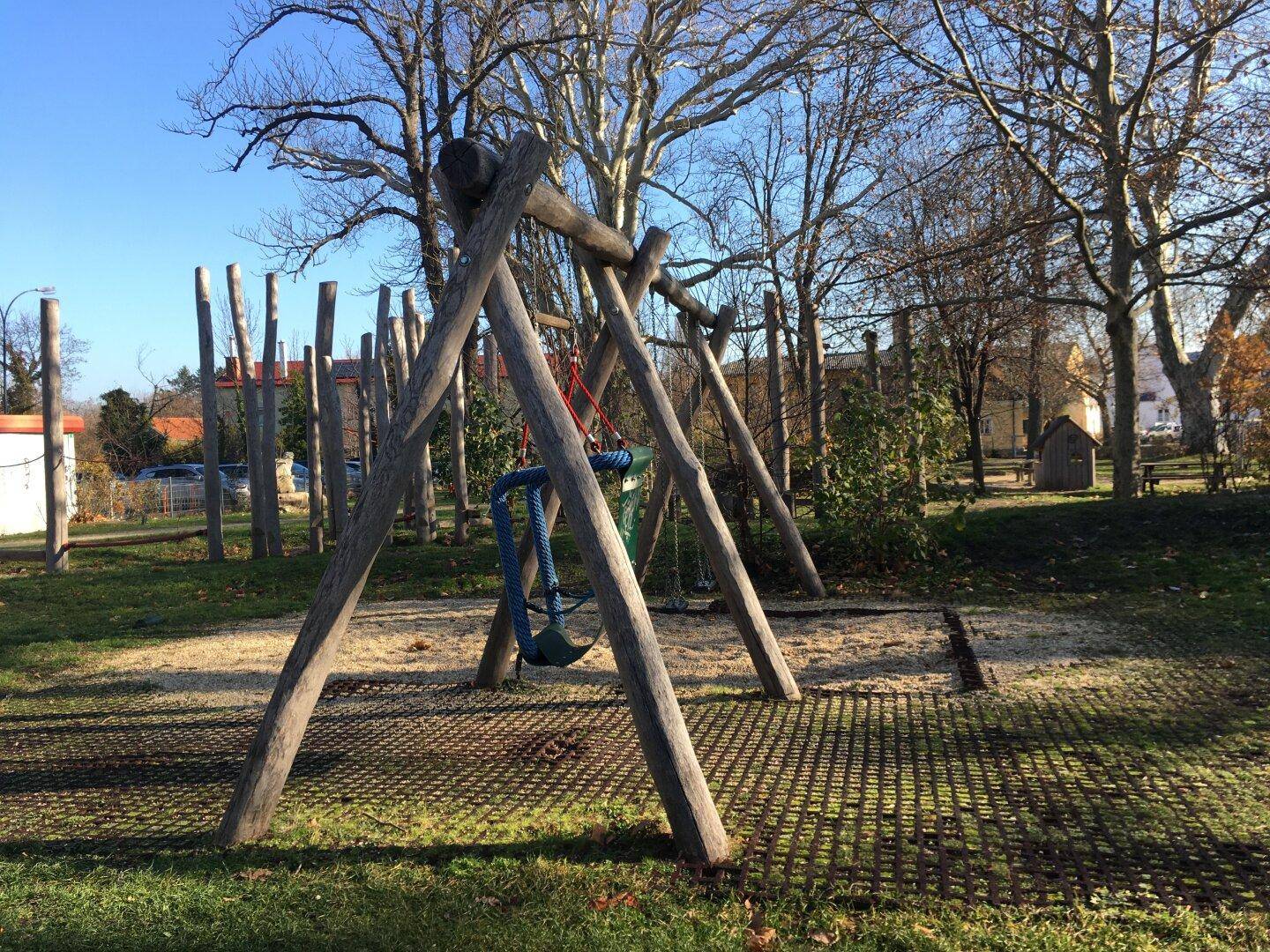 Öffentlicher Spielplatz mit Holzschaukeln und einem Sandbereich unter Bäumen.