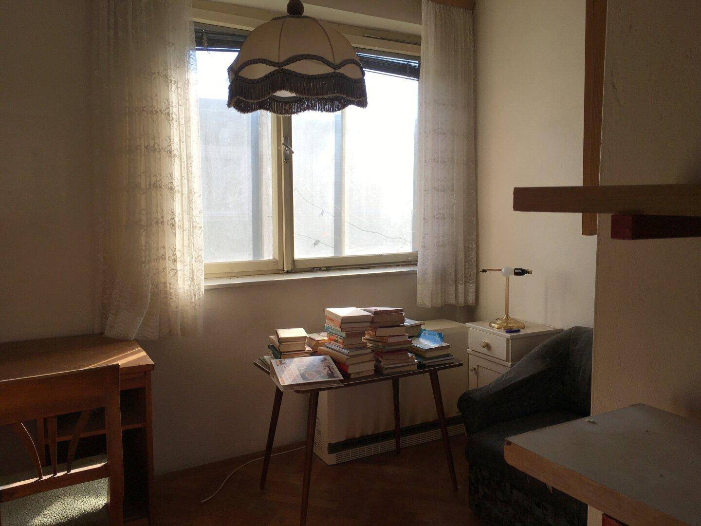 Helles Zimmer mit großem Fenster, Vorhängen und einem kleinen Tisch mit Büchern.