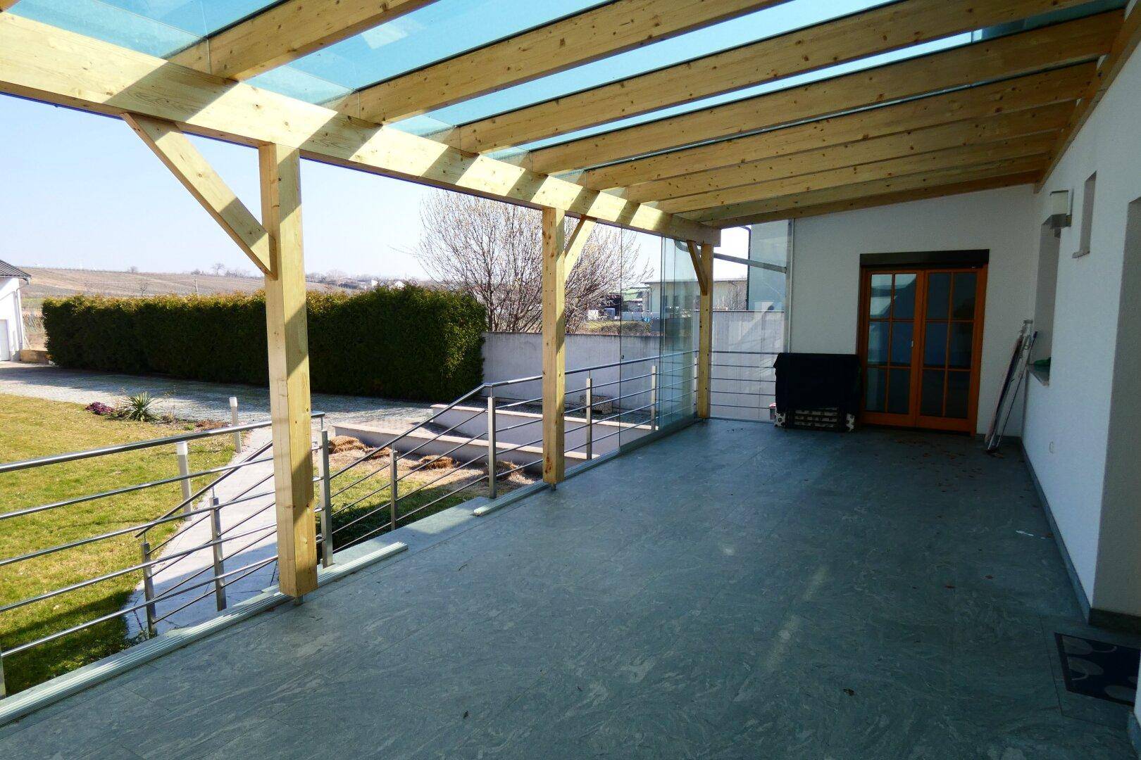 Geräumige überdachte Terrasse mit Holzbalkenkonstruktion, Fliesenboden und Blick ins Grüne.