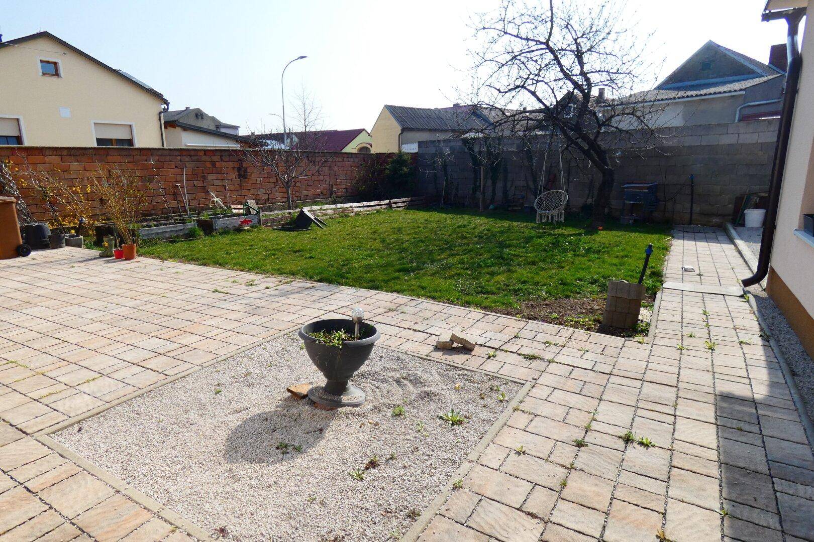 Sonniger Gartenbereich mit gepflasterter Terrasse, Rasenfläche und Kiesbeet.