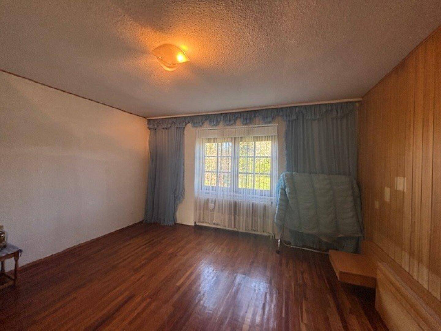 Helles Zimmer mit Holzpaneelen, Parkettboden und einem Fenster mit blauen Vorhängen.