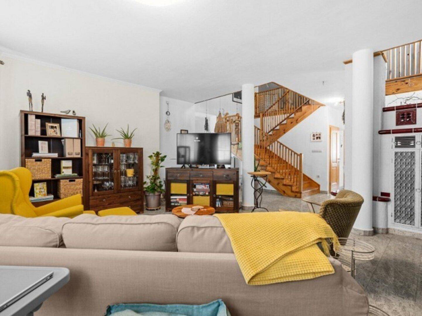 Gemütliches Wohnzimmer mit Sofa, Fernseher und Blick auf die offene Treppe und den Kachelofen.