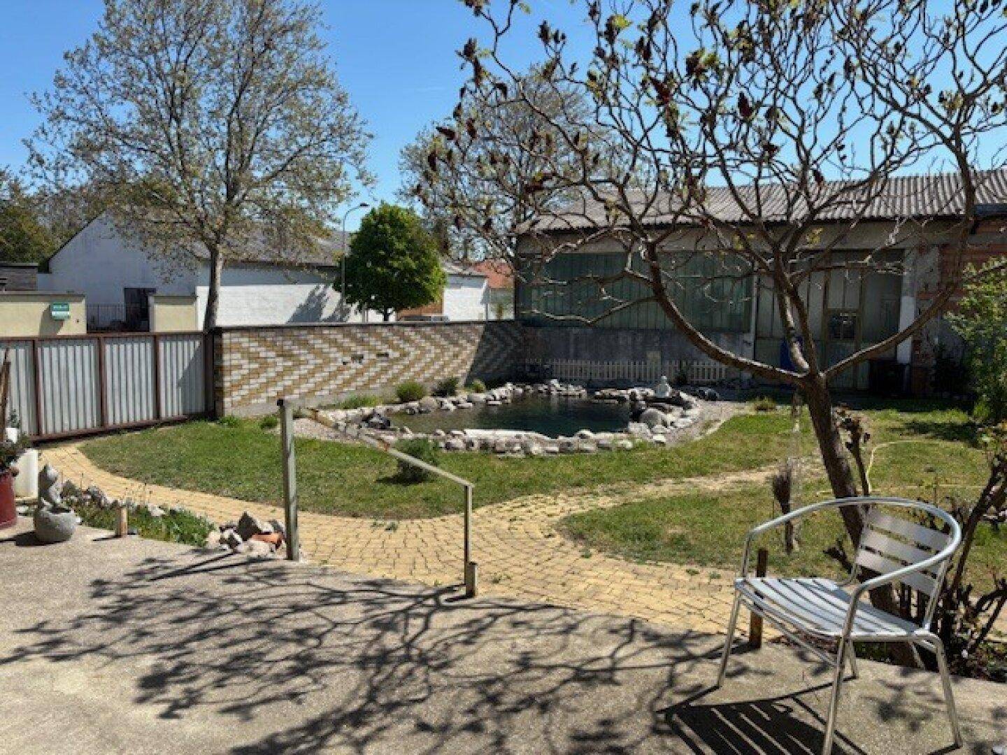 Sonniger Garten mit Teich, gepflasterter Terrasse und Sitzgelegenheit.