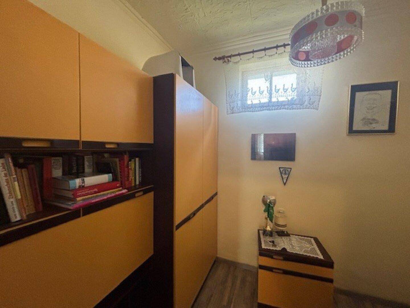 Kleiner Raum mit Bücherregal, Schränken und Fenster, das Tageslicht hereinlässt.