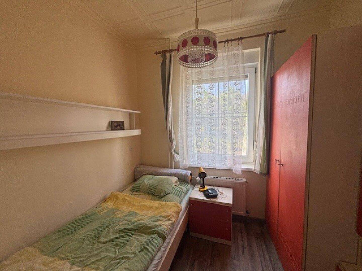 Schlafzimmer mit Einzelbett, rotem Kleiderschrank und Fenster mit Blick ins Grüne.