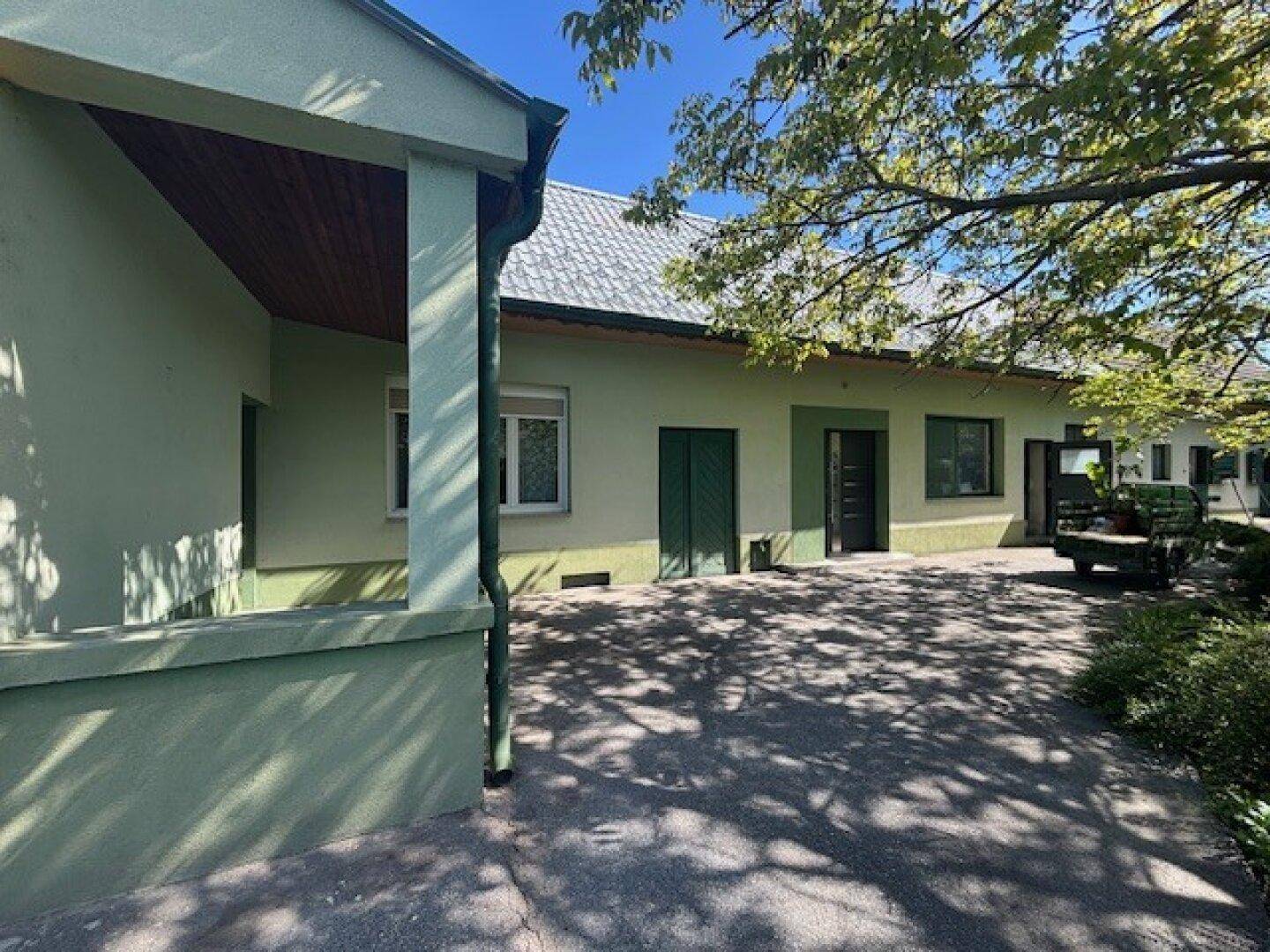 Seitenansicht des Hauses mit gepflastertem Bereich und Schattenwurf von Bäumen an einem sonnigen Tag.