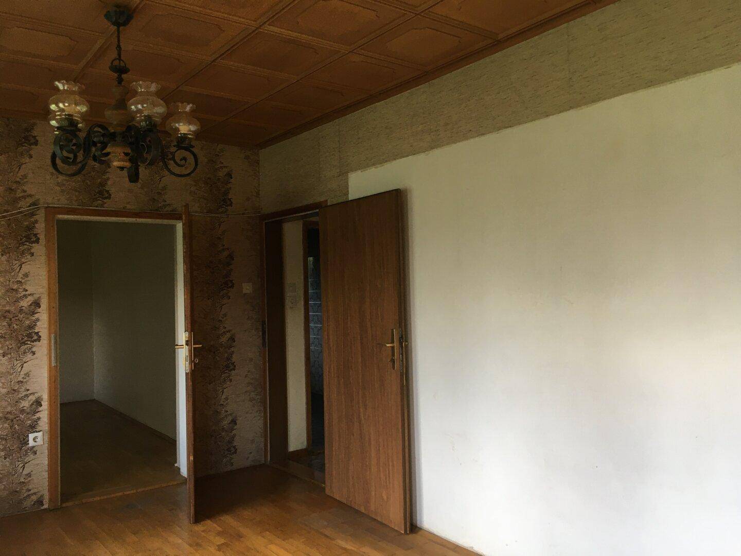Innenraum mit gemusterter Tapete, Holzdecke und einem Kronleuchter, bereit zur Renovierung.