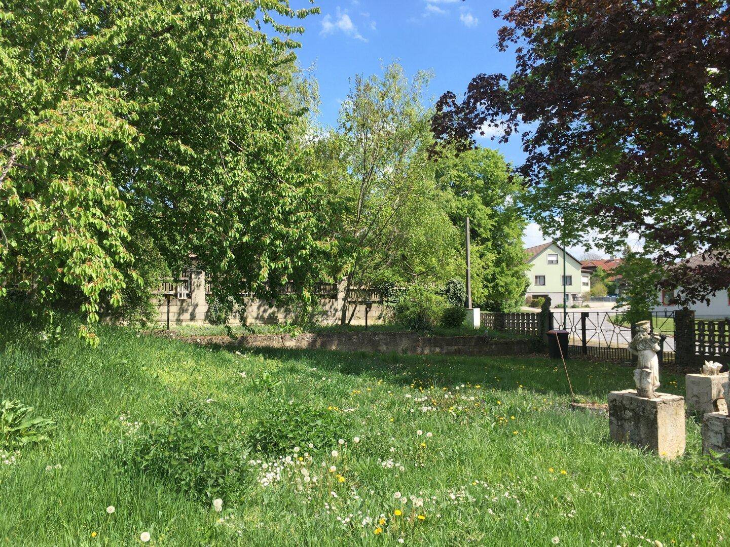 Sonniger Gartenbereich mit üppigem Grün, alten Bäumen und dekorativen Statuen im Freien.