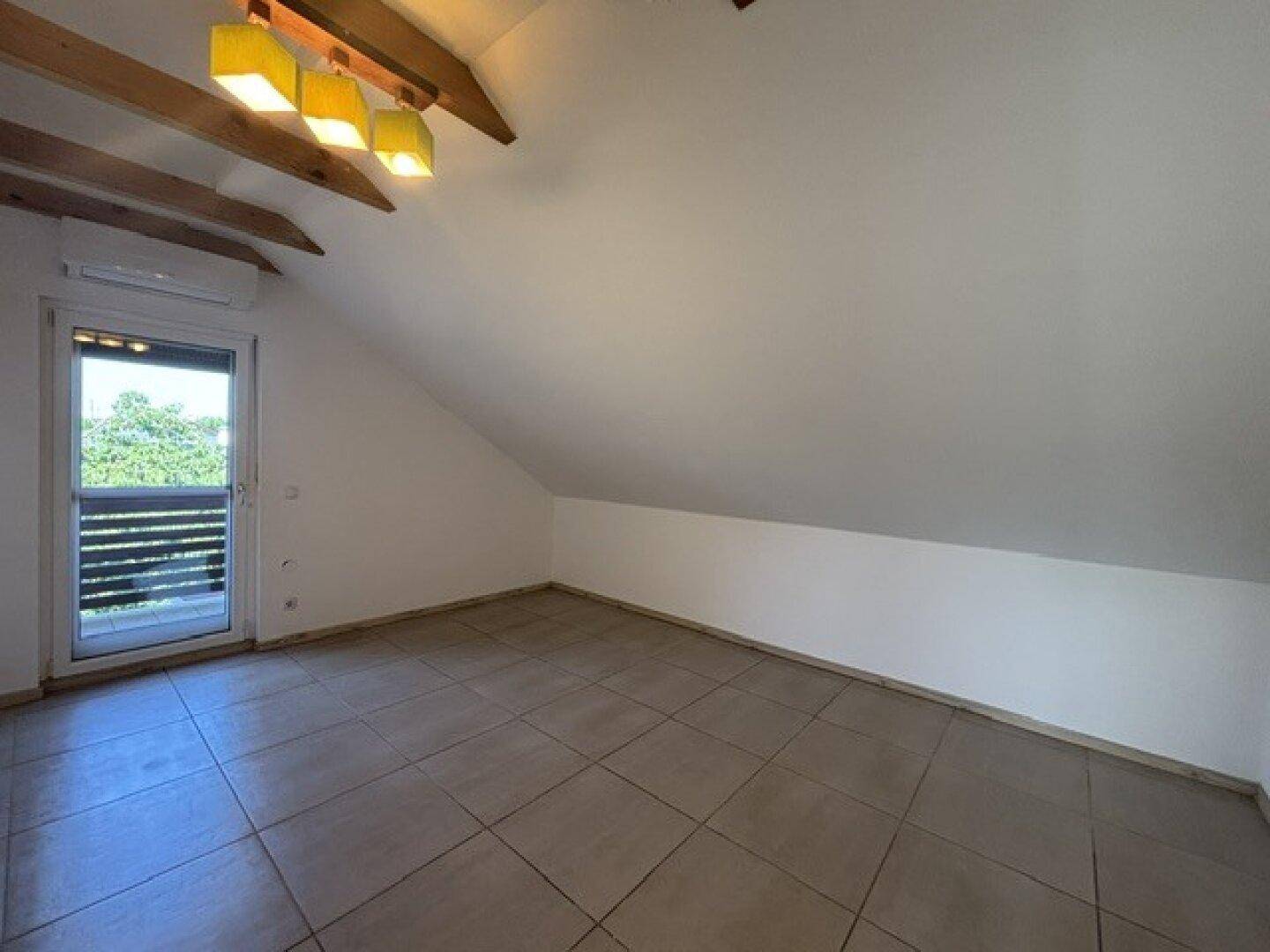 Helles Dachgeschosszimmer mit sichtbaren Holzbalken, Fliesenboden und Balkonzugang.