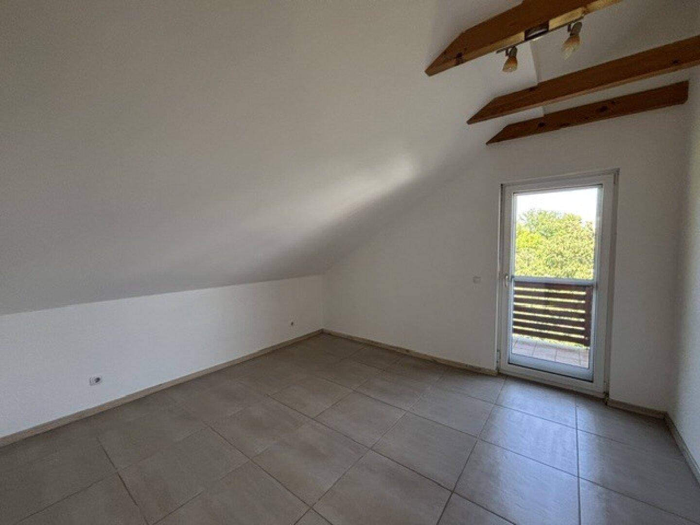Dachgeschosszimmer mit schrägen Wänden, Fliesenboden und Zugang zu einem kleinen Balkon.