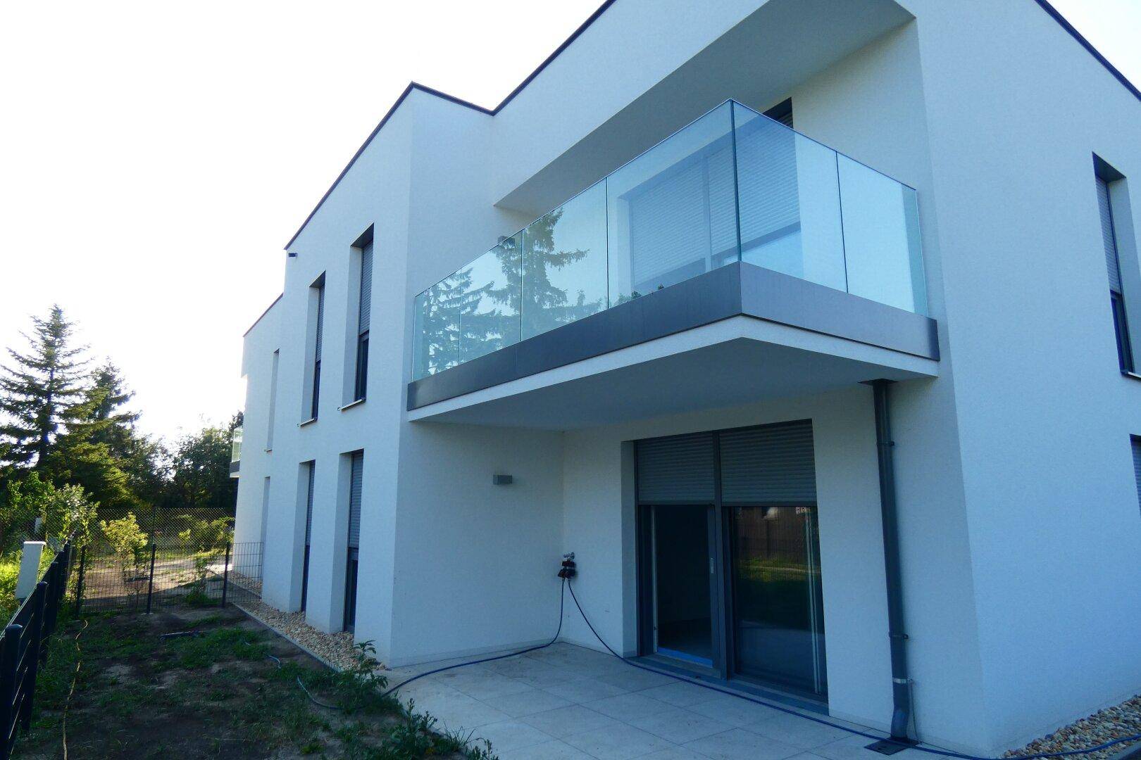 Seitenansicht des modernen Gebäudes mit Glasbalkon und einer kleinen Terrasse im Erdgeschoss.