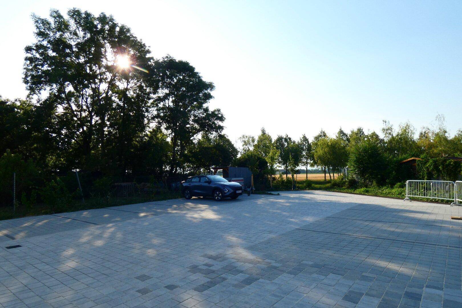 Großer gepflasterter Parkplatz im Freien, umgeben von Bäumen und mit Blick auf ein Feld in der Ferne.