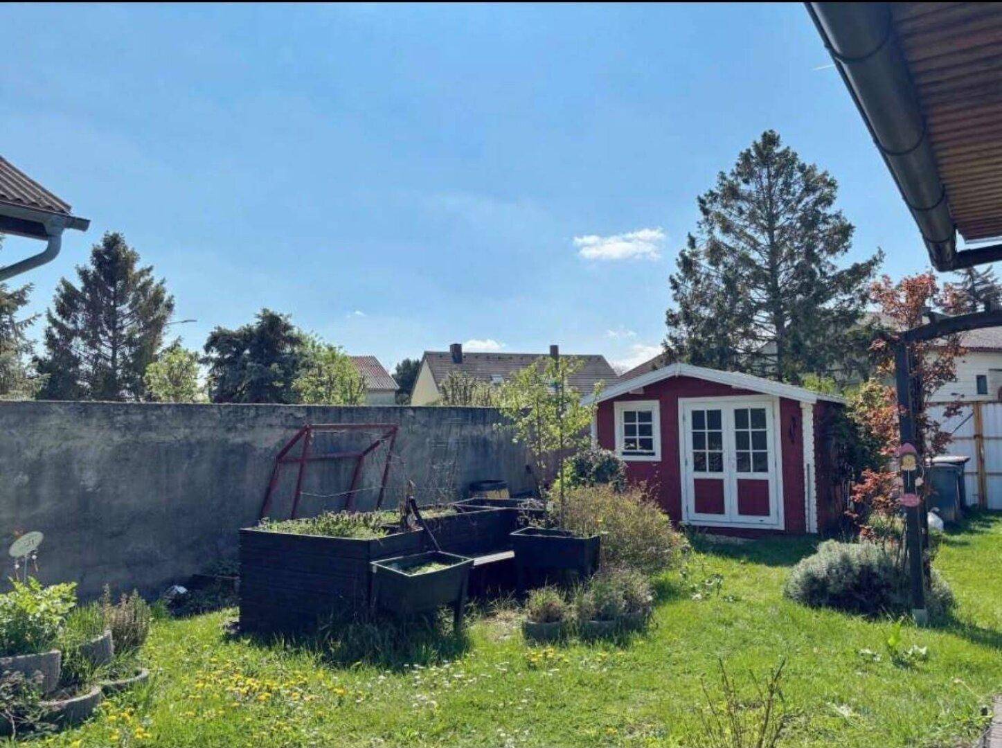 Grüner Gartenbereich mit einem roten Gartenhaus und Hochbeeten, umgeben von einer Mauer.
