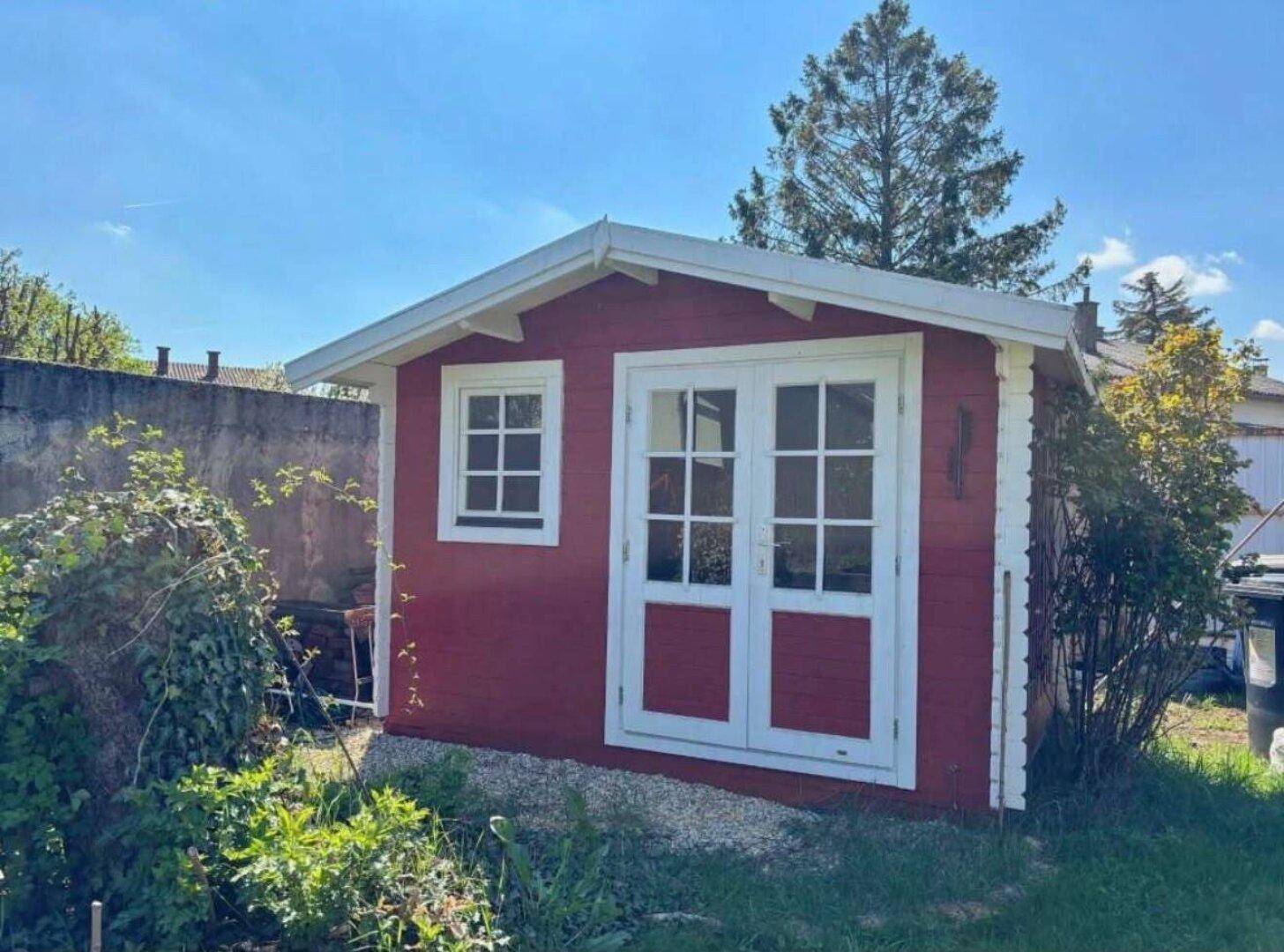 Charmantes rotes Gartenhaus mit weißen Türen und Fenstern, eingebettet in den grünen Garten.