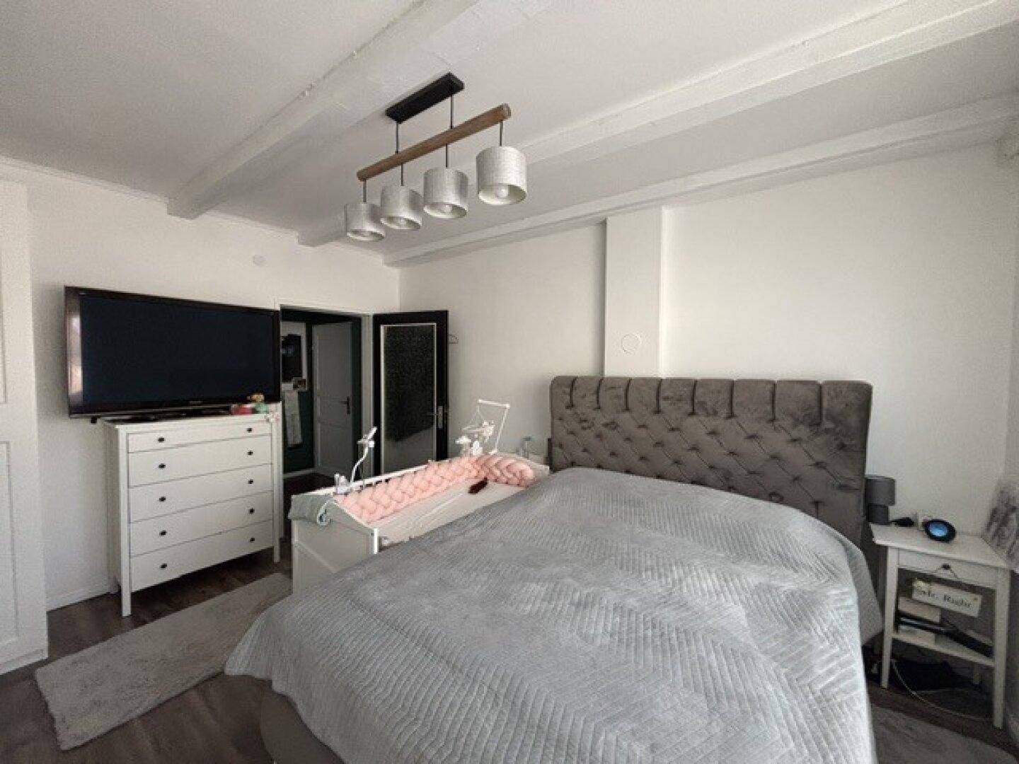 Gemütliches Schlafzimmer mit großem Bett, Babybett und Fernseher, ergänzt durch sichtbare Deckenbalken.