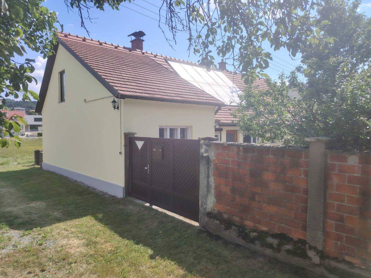 Seitenansicht eines gelben Hauses mit braunem Tor und Ziegelmauer im Grünen.
