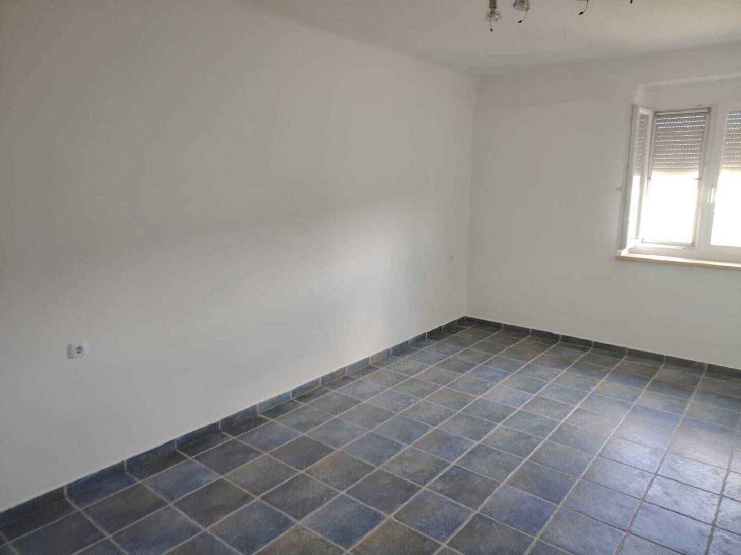 Heller Raum mit blauen Bodenfliesen und einem Fenster mit Rollladen.