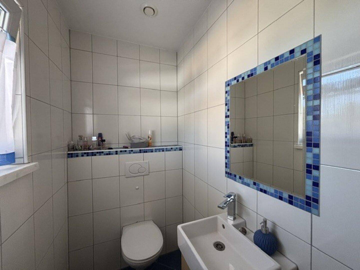 Modernes Gäste-WC mit weißen Fliesen, kleinem Waschbecken und Mosaikspiegel.
