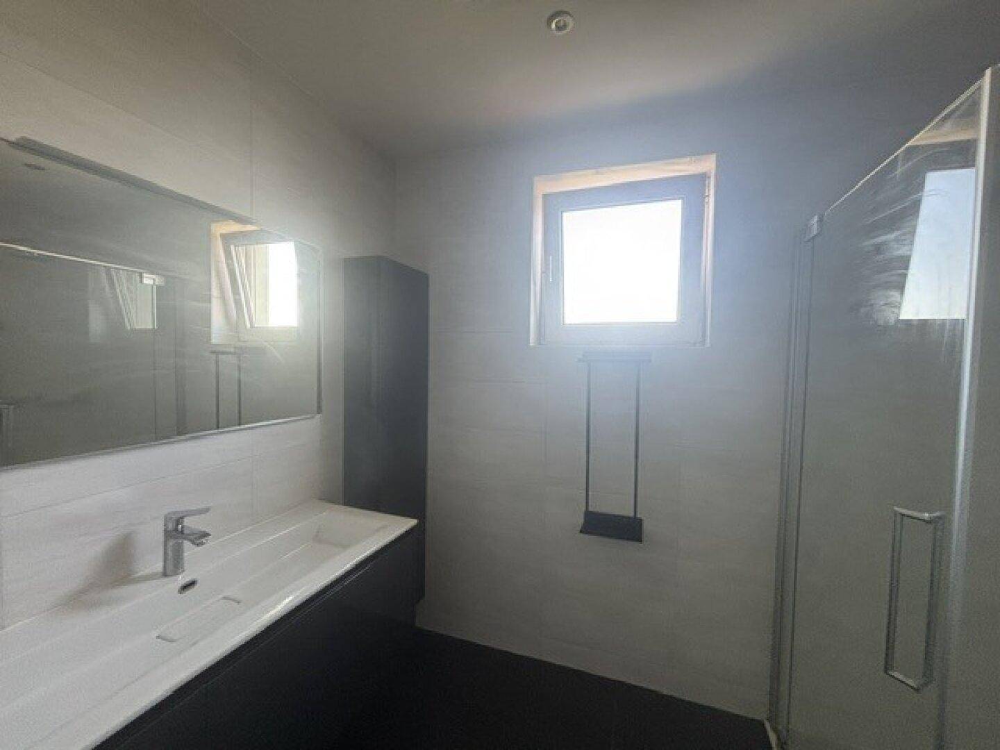 Modernes Badezimmer mit Einzelwaschbecken, großer Spiegelfläche, Duschkabine und Fenster.