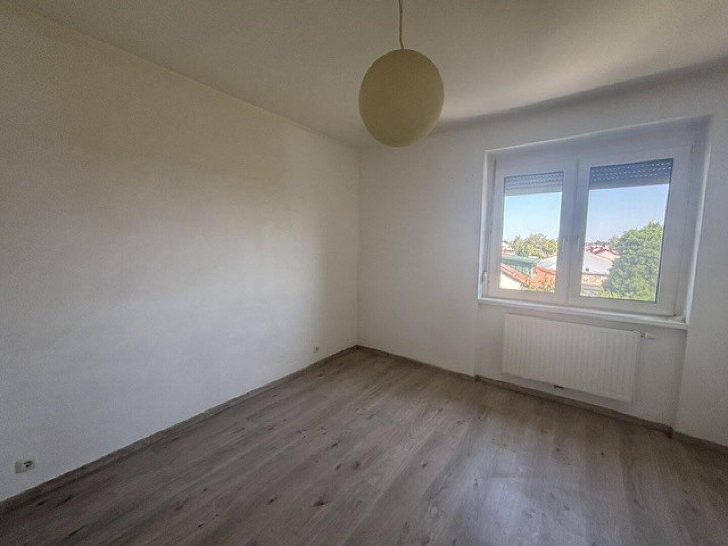 Helles Schlafzimmer mit Laminatboden und einem Fenster, das Tageslicht hereinlässt.