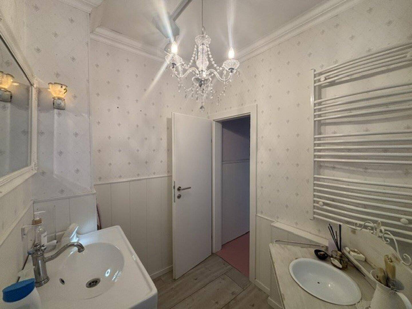 Helles Badezimmer mit zwei Waschbecken, Kristallleuchter und beheiztem Handtuchhalter.