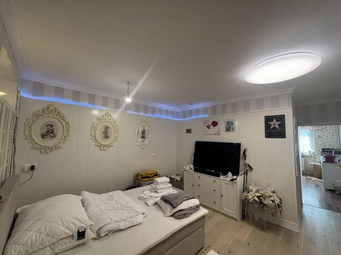 Schlafzimmer mit hellem Holzboden, indirekter Beleuchtung und einem Fernseher an der Wand.