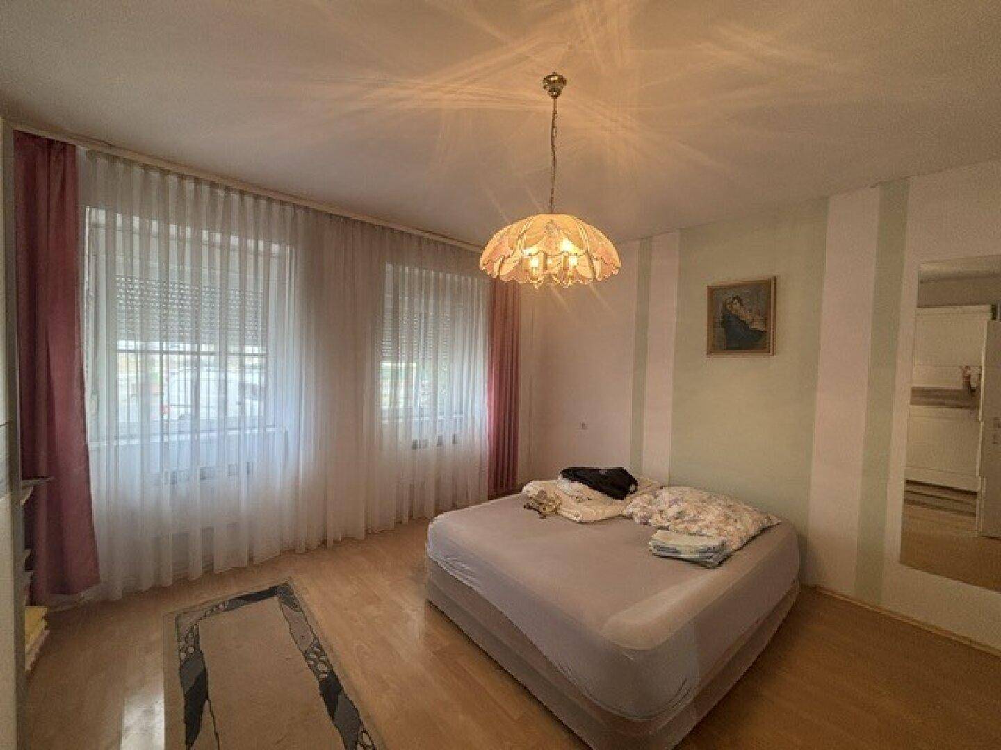 Schlafzimmer mit Holzboden, zwei Fenstern und einer dekorativen Deckenleuchte.