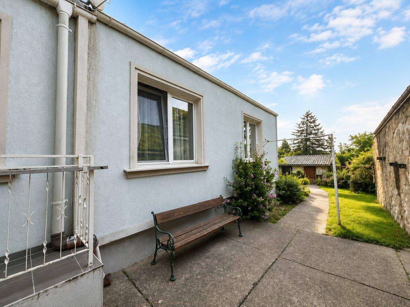 Seitenansicht des Hauses mit Fenster, einer Holzbank und einem gepflasterten Weg zum Garten.