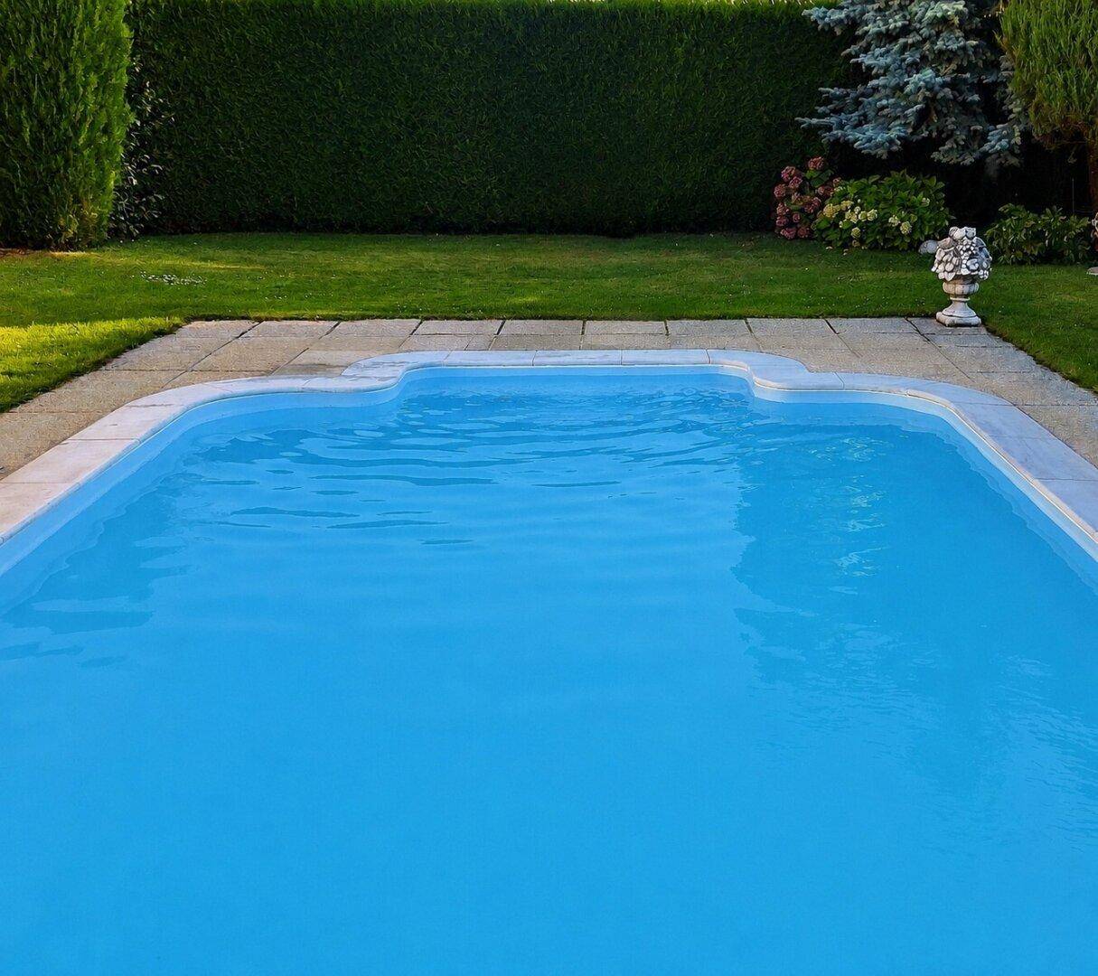 Blauer Swimmingpool im Garten, umgeben von einer gepflegten Hecke und Rasenfläche.