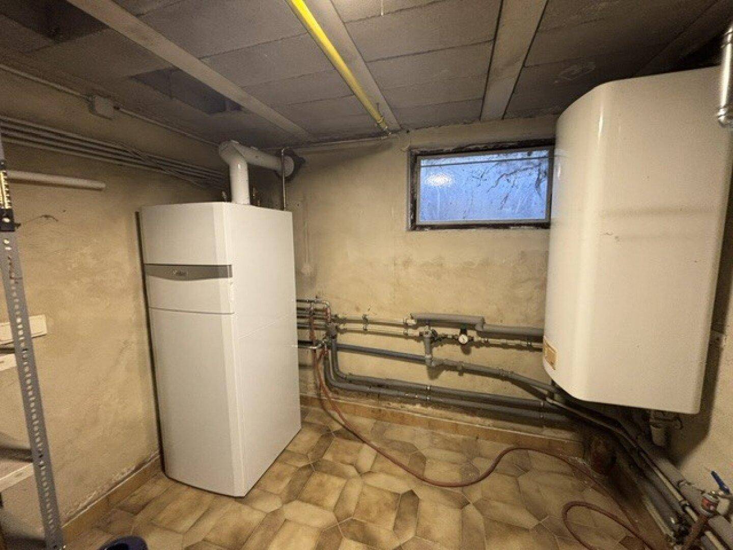 Technikraum im Keller mit Heizungsanlage, Warmwasserspeicher und sichtbaren Leitungen.