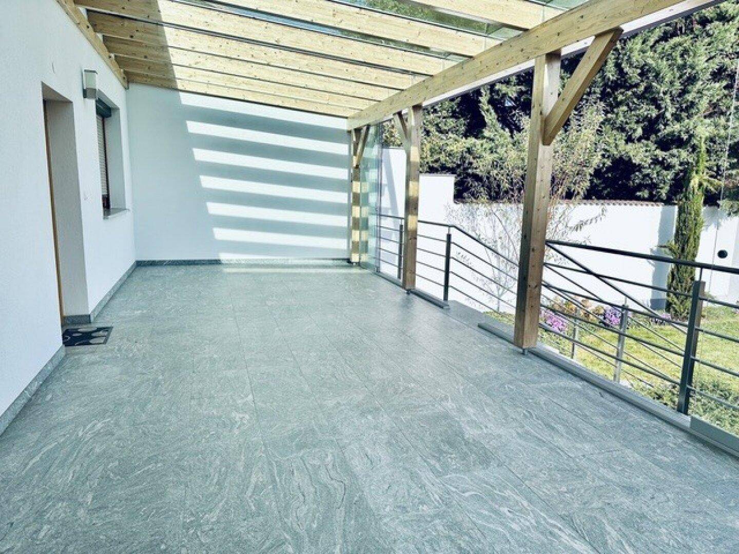 Helle, überdachte Terrasse mit modernen Fliesen und einer robusten Holz-Glas-Konstruktion, die viel Licht spendet.