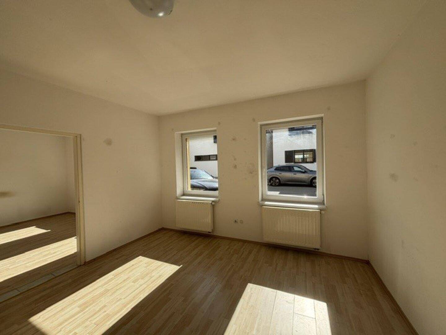 Leeres Wohnzimmer mit Laminatboden, Heizkörpern unter den Fenstern und natürlichem Lichteinfall.