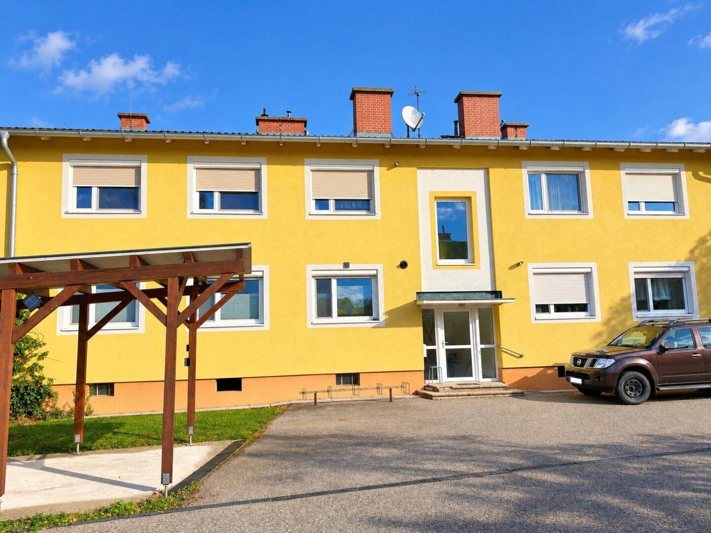 Gelbes Mehrfamilienhaus mit Carport und Parkplätzen vor dem Eingangsbereich.