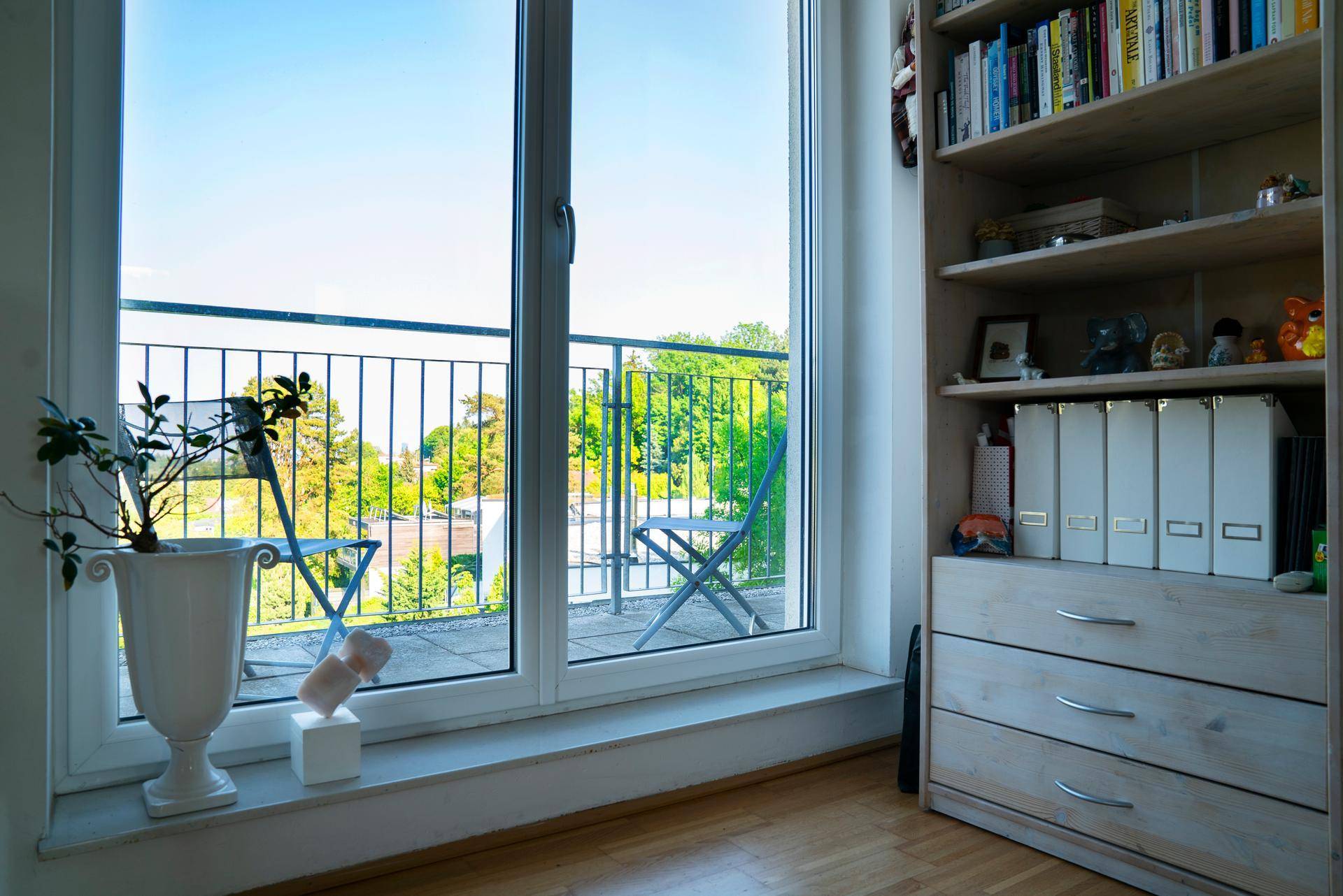 Heller Raum mit Bücherregal und Fenster mit Blick auf einen Balkon und die grüne Umgebung.