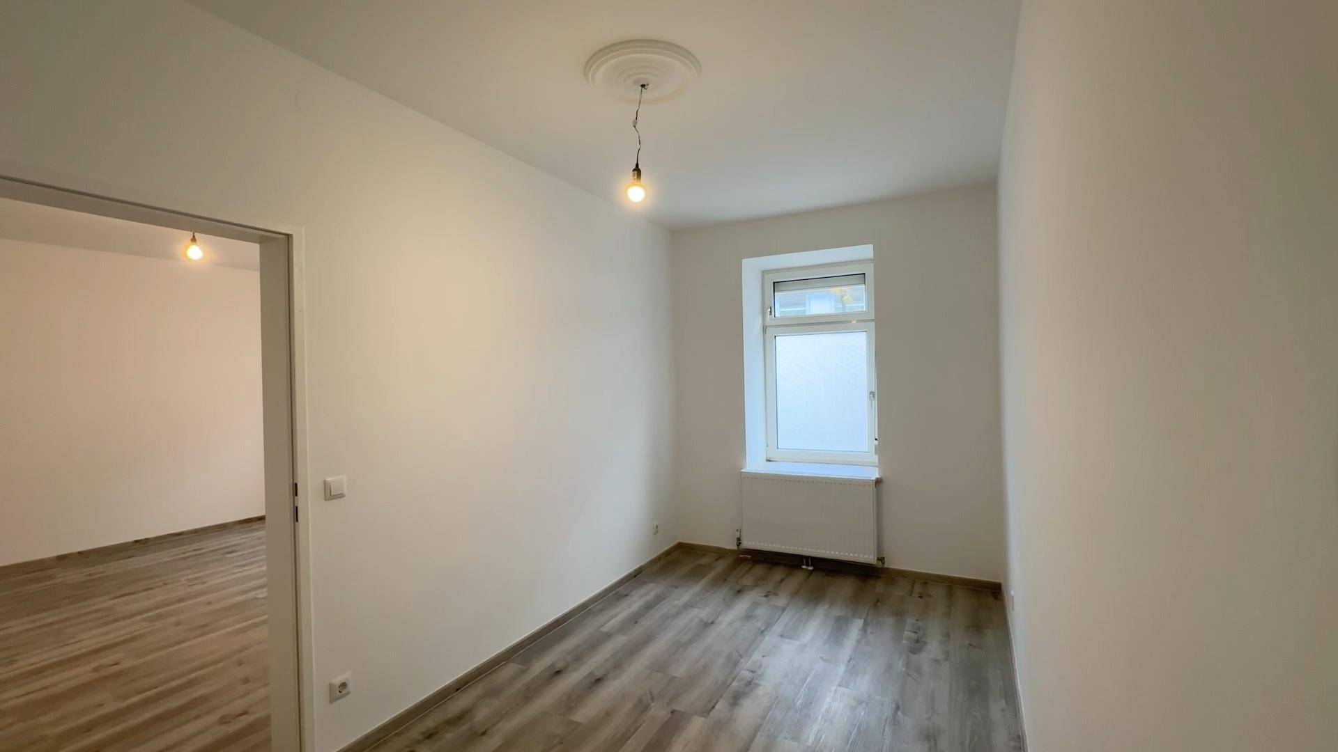Helles Zimmer mit Laminatboden und einem Fenster, verbunden mit einem weiteren Raum.