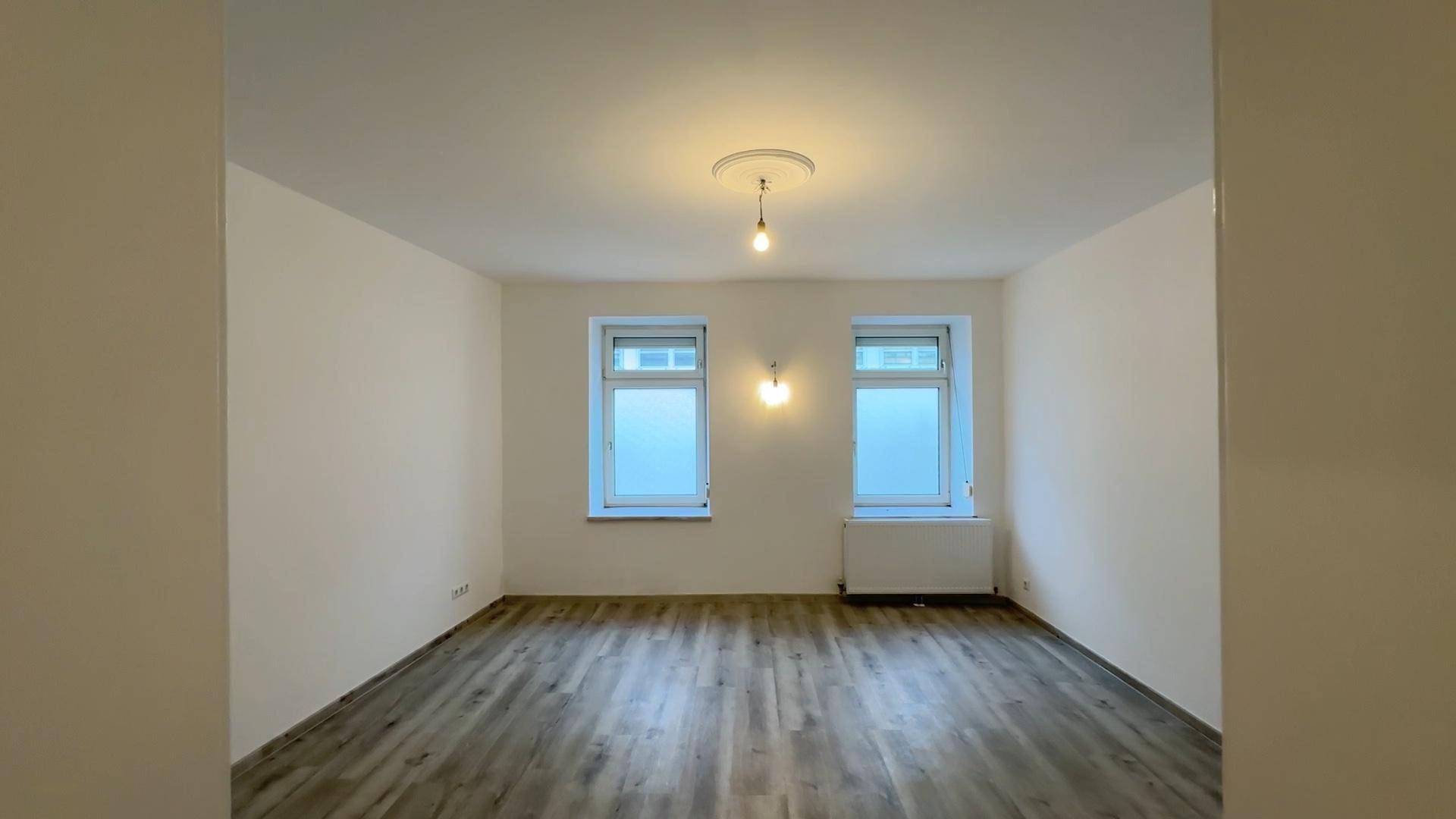 Geräumiges Zimmer mit hellem Laminatboden und zwei Fenstern, ideal für Wohn- oder Schlafbereich.