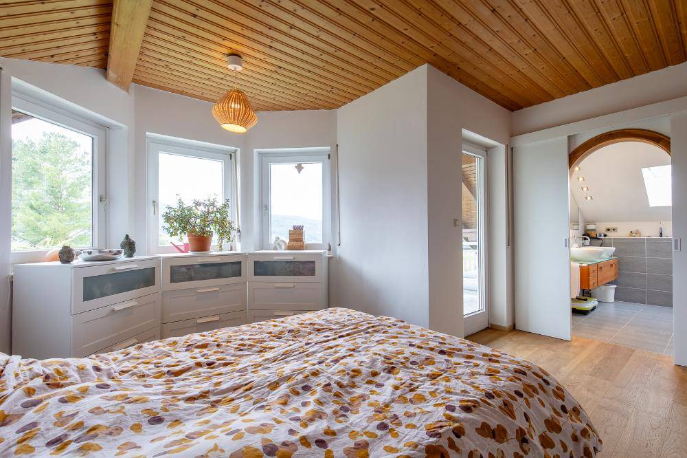 Helles Schlafzimmer mit Holzbalkendecke, großen Fenstern und direktem Zugang zum Badezimmer.
